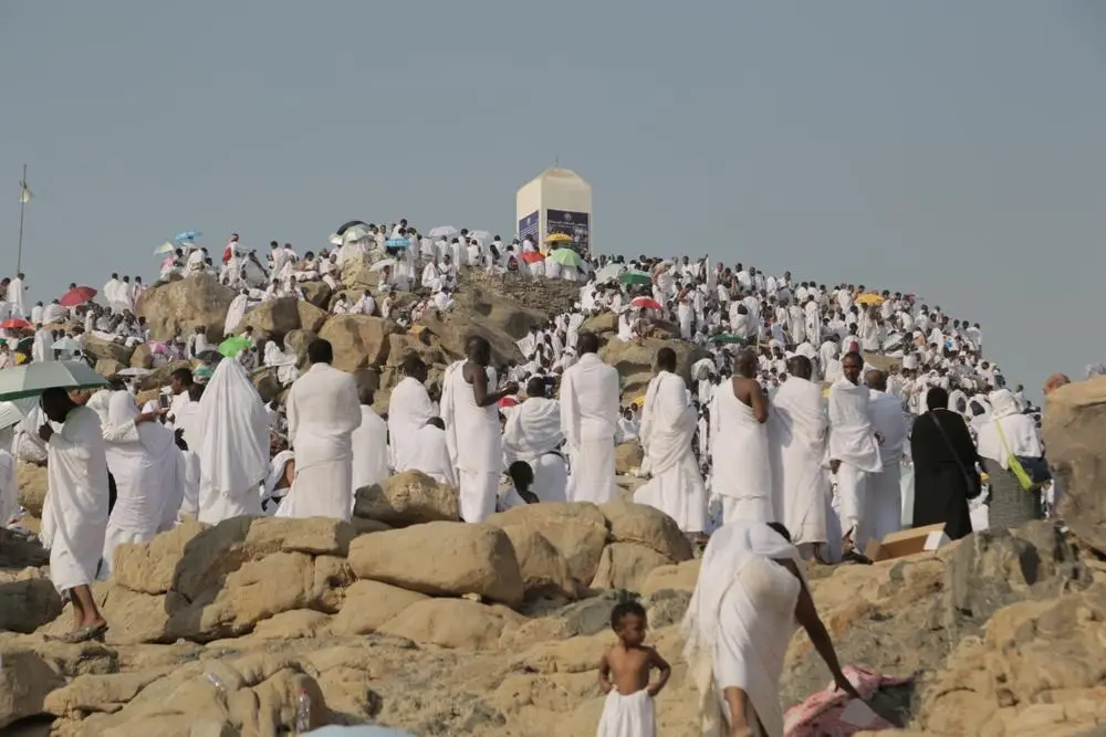 Pilgrims in white on the rocks of Jabal ar-Rahmah at Arafah on the day, umbrellas scattered through the crowd, the white marker pillar at the summit against a pale sky.