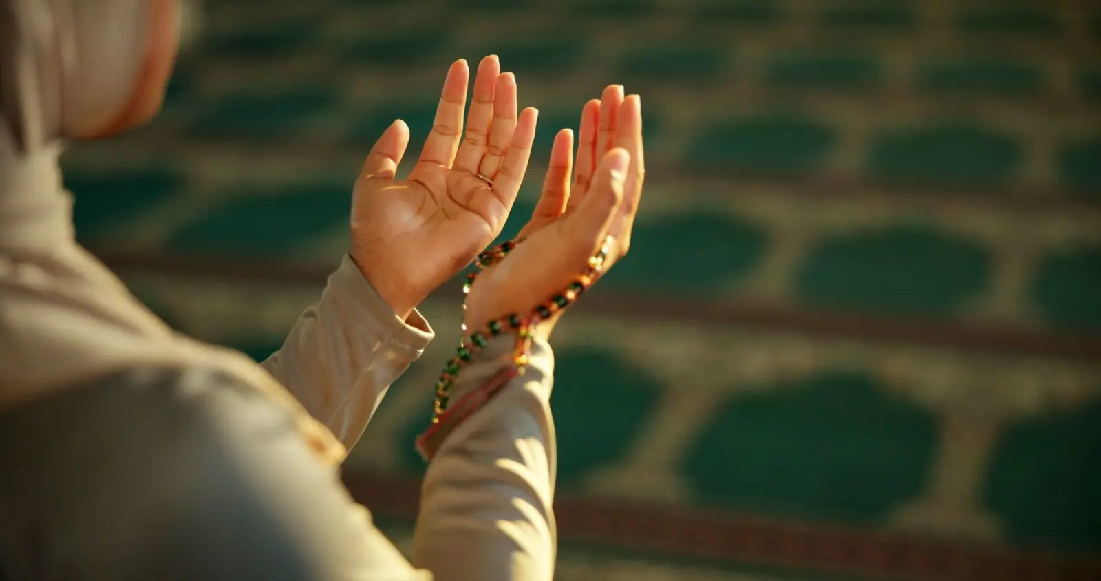Two open hands raised in du‘a, a string of prayer beads wound around one wrist, the green and red of a mosque carpet blurred behind them.