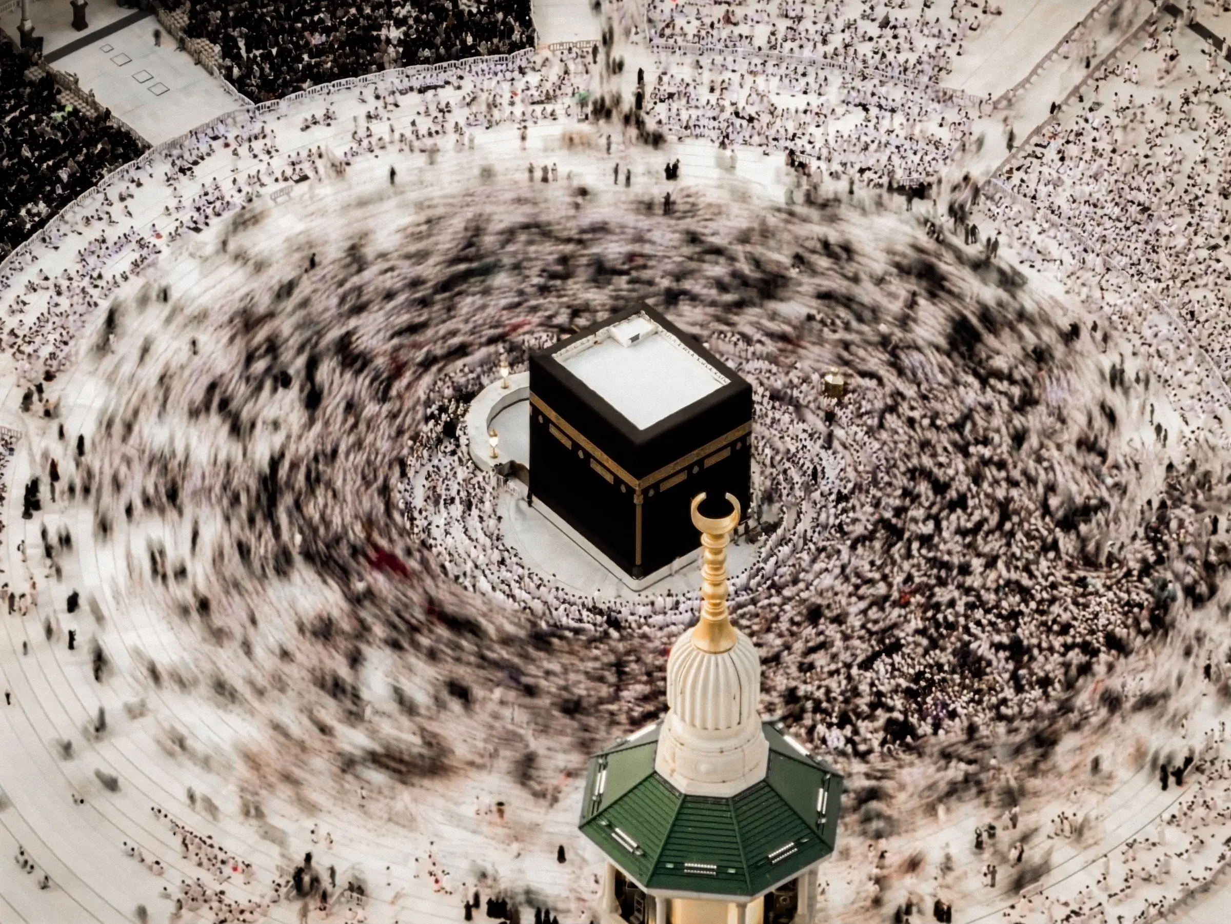 The Ka‘bah seen from directly above in daylight, the crowd circling it drawn by a long exposure into a single blurred spiral, with the green and gold cap of a minaret rising in the foreground.