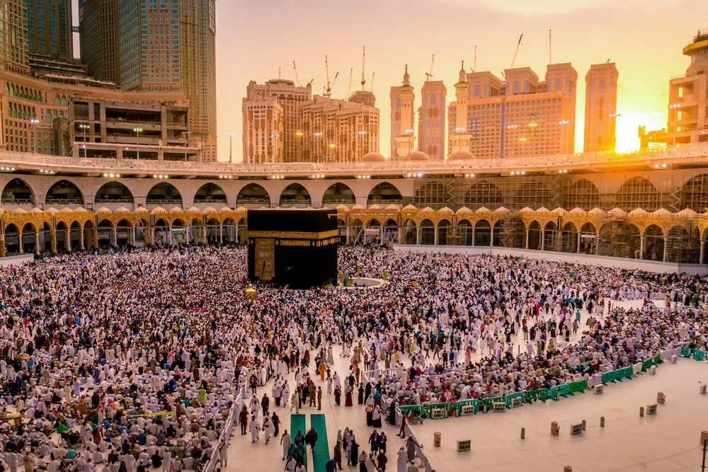The Grand Mosque at sunset from above the courtyard, the Ka‘bah at the centre of a dense crowd, the towers of the city catching the last light behind the arcade.