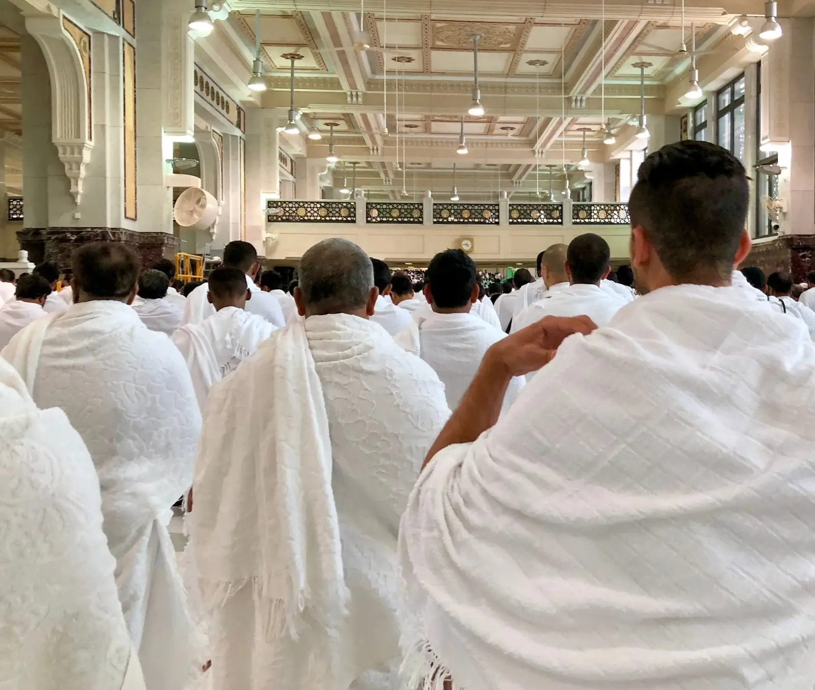 Men in ihram seated close together in a mosque hall, all seen from behind, the white cloth over one shoulder and the bare shoulder showing.
