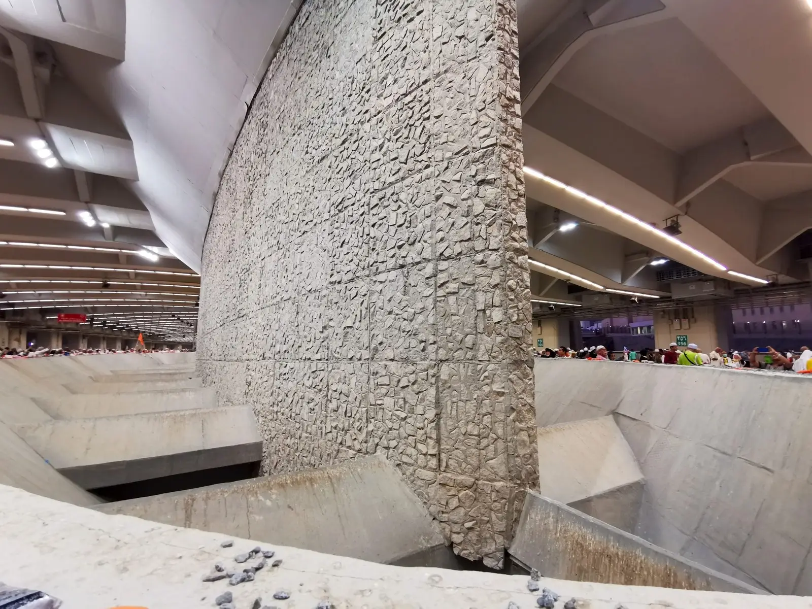 One of the Jamarat pillars inside the multi-storey bridge at Mina: a tall wall of rough stone dropping into a concrete basin, with pilgrims at the rail on the level above.