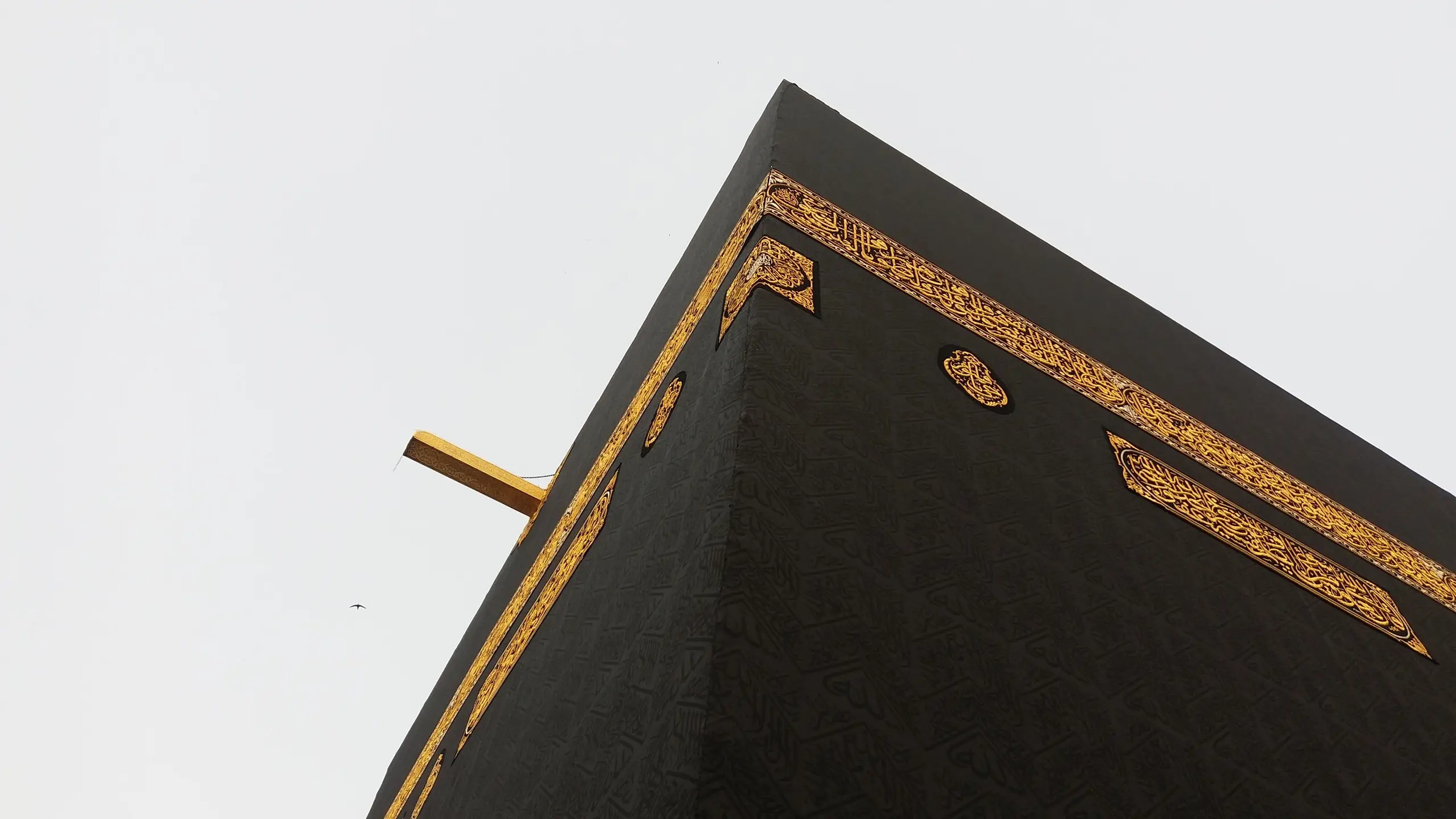 The upper corner of the Ka‘bah seen from below against a pale, overcast sky, the black kiswah rising across the frame with its band of gold-embroidered calligraphy, and the golden waterspout projecting from the roof edge.