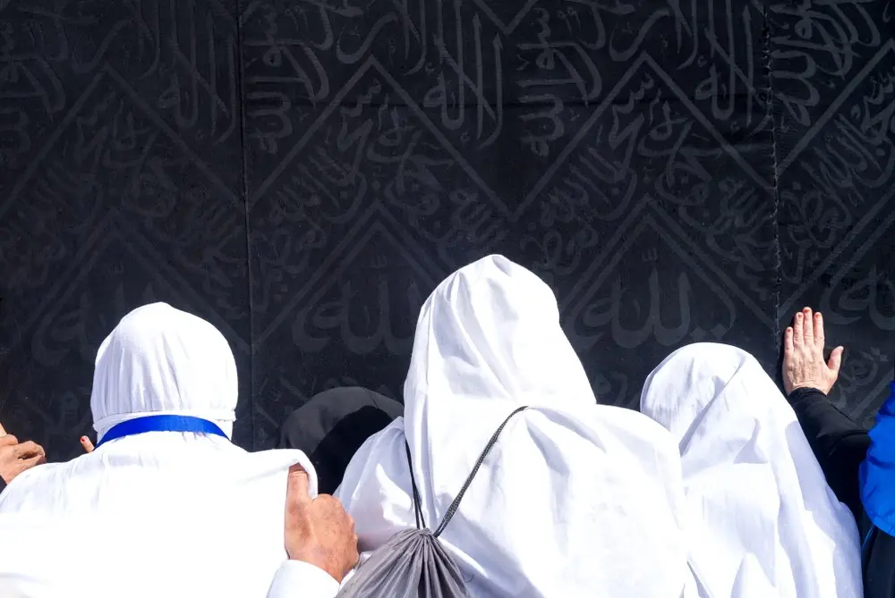 Pilgrims pressed against the kiswah with their hands flat on the black cloth, the silver embroidery of its calligraphy filling the frame above them.