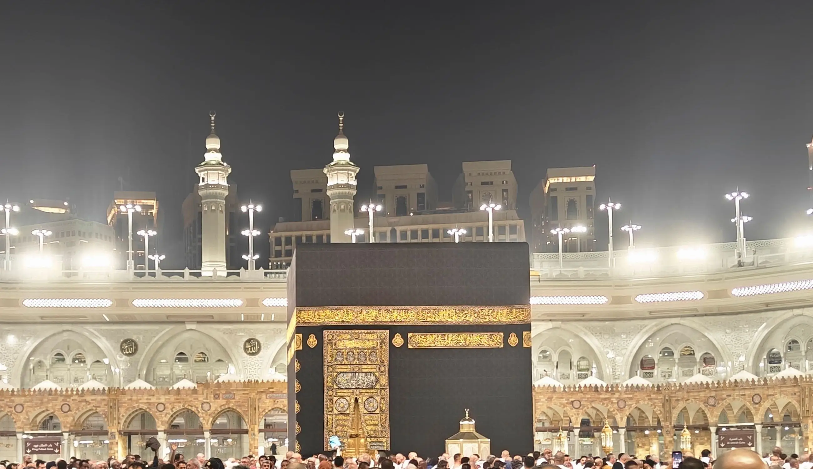 The Ka‘bah floodlit at night, the gold-embroidered door and band of the kiswah catching the light, minarets and the arcades of the Grand Mosque behind, a dense crowd at its foot.