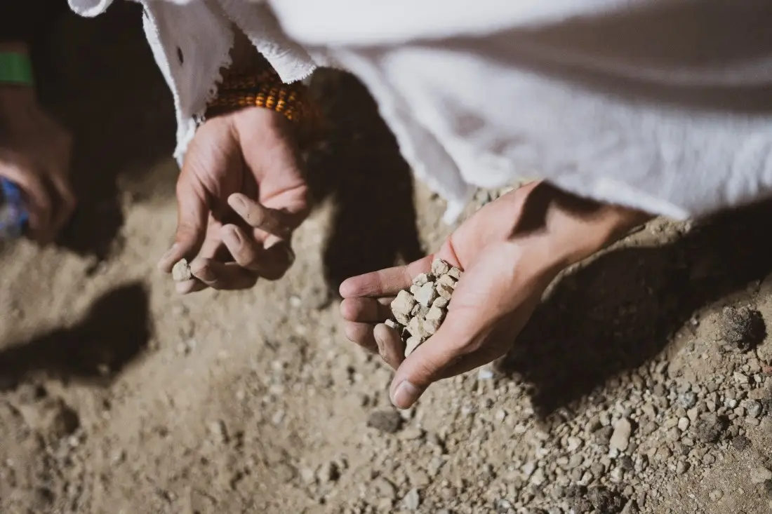 Two hands in Ihram close over the open ground at Muzdalifah, one cupping a small pile of pebbles gathered for the Jamarat and the other reaching for more.