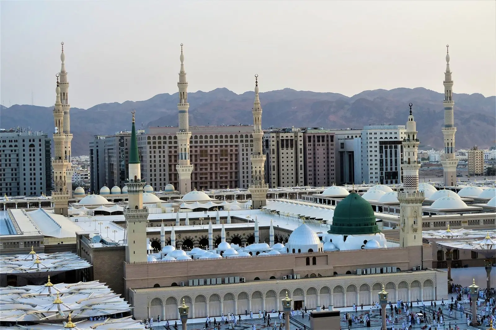 The Prophet’s Mosque in Madinah from above: the green dome and pale minarets among the white roofs of the prayer halls, bare mountains behind.
