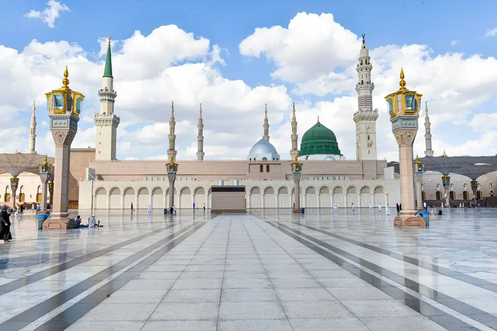 The marble courtyard of the Prophet’s Mosque in daylight, almost empty, the green dome and the minarets rising beyond the arcade, folded canopies on their columns to either side.