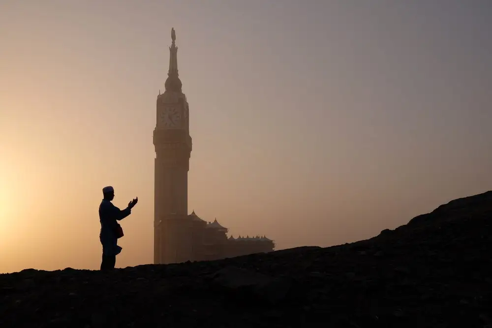 A man standing on a rocky ridge at dusk with his hands raised in du‘a, in silhouette against an orange sky, the clock tower of Makkah hazy behind him.